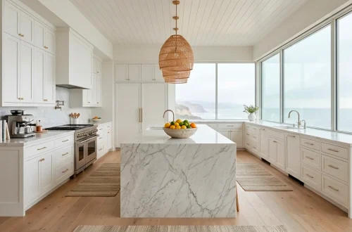 Wide horizontal view of a bright coastal kitchen with white cabinets, marble island, and ocean views through large windows.