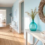 Wide landscape photo of a bright coastal entryway with white furniture and turquoise glass accents.