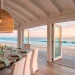 Wide panoramic view of an airy coastal dining room with white beams, rattan pendant, and ocean views at sunset.