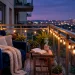 Wide shot of a cozy small balcony reading nook with a navy armchair, fairy lights, and city views at dusk.