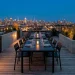Wide industrial patio banner featuring a modern black metal dining set on a city rooftop at dusk.