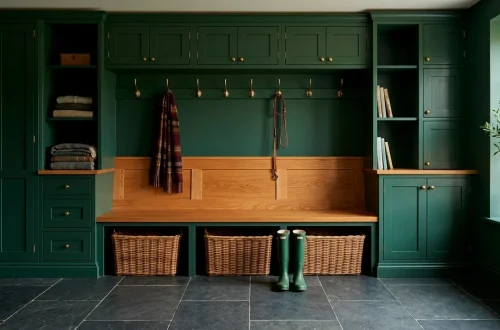 Wide landscape of a moody forest green mudroom with brass hooks, wood bench, and organized storage.