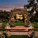 Wide view of a modern outdoor wedding stage decorated with lush florals and a glowing neon sign at dusk