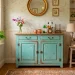 A distressed teal home bar cabinet styled in a sunlit shabby chic living room with crystal glasses and liquor bottles.