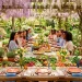 Sunlit outdoor Easter dining table setting with fresh spring vegetables, potted herbs, and colored glassware.