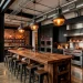 Wide view of a modern industrial kitchen featuring exposed red brick walls, matte black metal pipes, and a reclaimed wood island.