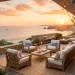 Beautiful coastal outdoor patio at golden hour with teak furniture, white cushions, and string lights overlooking the ocean.