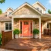 Bright coral front door on a summer porch with green ferns and modern concrete planters in warm sunlight.
