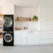 Sleek minimalist small laundry room featuring stacked appliances, custom wooden shelving, and clean white cabinetry.
