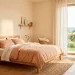 A wide view of a soft minimalist bedroom featuring a natural oak bed, creamy walls, and an olive tree illuminated by spring sunlight.