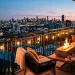 A cozy, warm balcony at twilight featuring a blazing tabletop fire pit, string lights, and chunky knit blankets.
