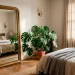 Large floor mirror in a small sunlit bedroom reflecting a cozy bed and plants to create depth