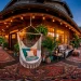 Wide bohemian wrap-around porch featuring a white macrame hanging hammock, layered vintage rugs, and lush potted plants.