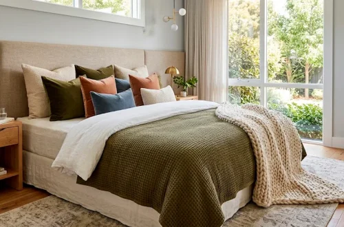 A perfectly layered modern bed featuring linen sheets, a folded white duvet, an olive-green quilt, and a chunky knit throw blanket.