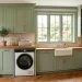 Wide view of a modern laundry room featuring sage green built-in cabinets and sleek washing machines under bright sunlight.