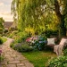 Wide view of a romantic garden reading nook featuring a vintage wrought iron bench beneath a weeping willow tree.