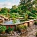Beautiful wooden above-ground pond with a waterfall on a backyard patio surrounded by lush green plants and river stones.