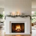 Minimalist white Christmas fireplace decor featuring frosted pine garlands, silver candles, and white ceramic houses.