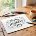 Aesthetic flat lay of a hand-lettered Mother's Day card in progress on a wooden desk with brush pens and tea