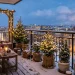 A cozy snow-covered urban balcony with potted evergreens wrapped in burlap and glowing string lights for winter.