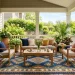 A sunlit summer porch featuring a vibrant geometric outdoor rug under modern teak patio furniture.