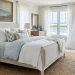 Wide view of a coastal grandmother guest room featuring breezy white linens, woven rattan rug, and vintage wood nightstand.